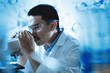 © KANGWANS - A men wearing a lab coat and safety glasses is looking through a microscope.