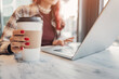 © EdNurg - Woman holding a disposable coffee cup while using a laptop on a marble table, working remotely in a cafe during the day