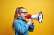 © miss irine - Young girl wearing glasses and blue sweater speaks loudly into red and white megaphone. Kid makes announcement against plain yellow background, copy space available for text.