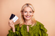 © deagreez - Happy mature woman with blonde hair holds credit cards smiling against beige background wearing a green blouse