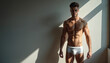© Viktor - Fit man stands by window in white briefs. Sunlight creates shadows on wall. Athletic male body, six pack abs, strong arms, healthy physique, attractive male model poses in studio. Fashion underwear.