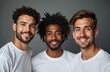 © Viktor - Three diverse men smile together in white t-shirts on a gray studio background. They represent multicultural male friendship and skin positivity with natural grooming and clear complexions.