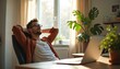 © Viktor - Young man rests at home office chair with hands behind head. He looks relaxed and happy while taking a break from computer work near potted plants by sunlit window, enjoying peaceful moment.