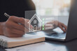 © Kerianna - Person planning real estate investment or home purchase, writing notes and using a laptop with a virtual house icon and checklist for property management or mortgage application tasks.