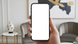 © MD Rashedul Islam - Hand holding mobile smartphone mockup with blank white screen in vertical portrait position against blurred luxury living room interior with contemporary furniture for app design display