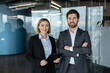 © Liubomir - Two smiling business professionals, a woman holding a clipboard and a man with crossed arms, standing confidently in a modern glass office portraying teamwork and success