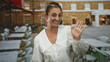 © Krakenimages.com - Woman smiling and waving hand on a restaurant terrace with tables and chairs visible, white blouse and earrings, frontal crop and direct eye contact; friendly greeting.