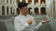 © Krakenimages.com - Young hispanic man in striped sweater pointing and presenting with hand in a historic university courtyard, showcasing focus and engagement.
