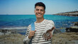 © Krakenimages.com - Young man with a striped shirt smiles warmly at the seaside, extending his hand towards the camera, with a turquoise sea in the background, capturing a friendly summer moment.