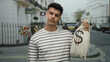 © Krakenimages.com - Young man on city street holding money bag symbolizing wealth and success with a confident expression in an urban outdoor setting.