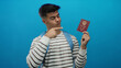 © Krakenimages.com - Young man with a blue background holding a chinese passport touches his head in thought, wearing a striped shirt and a blue backpack, implying travel and identification theme.