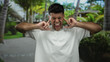 © Krakenimages.com - Young man outdoors in park covering ears with fingers looking frustrated amidst tropical backdrop.