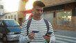 © Krakenimages.com - Young man outdoors on a city street holding smartphone and credit card wearing striped shirt and backpack captured in a vibrant urban environment.