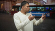 © Krakenimages.com - Man holding smartphone, tapping screen with furrowed brow while glancing at bus and city lights on busy street; digital frustration.