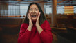 © Krakenimages.com - Young woman in red sweater expressing surprise in a modern indoor cafe setting with soft lighting, showcasing joy and excitement.