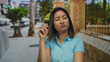 © Krakenimages.com - Young asian woman in blue shirt looks frustrated outdoors on a city street, surrounded by urban scenery and palm trees, conveying emotions of boredom and contemplation.