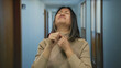 © Krakenimages.com - Young woman in hallway appearing sick holding throat wearing beige sweater in hotel setting with blurred background representing discomfort and health issues indoors.