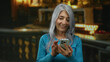 © Krakenimages.com - Senior woman with grey hair using a smartphone indoors in a church setting, smiling and wearing a blue sweater, with candles softly glowing in the background, capturing a serene moment.