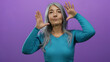 © Krakenimages.com - Senior woman with grey hair in a blue sweater poses against an isolated purple background, expressing with hands raised near her face.