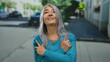 © Krakenimages.com - Senior woman with grey hair points with both hands while standing on an urban street in the outdoor setting, wearing a turquoise sweater and smiling softly in the bright daylight.