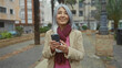 © Krakenimages.com - Senior woman with grey hair using smartphone outdoors on city street surrounded by urban buildings and palm trees, dressed warmly in scarf and coat on an overcast day.
