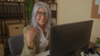 © Krakenimages.com - Senior grey-haired woman sitting in an office setting, engaged and gesturing with her hand while working at a desk with a computer, surrounded by shelves and decor.