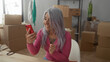 © Krakenimages.com - Senior woman with grey hair using smartphone in her new home living room, surrounded by moving boxes and unpacked items, expressing surprise and joy.