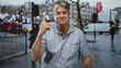 © Krakenimages.com - Middle aged man pointing index finger upward on busy wet street, smiling in short sleeve shirt and grey hair; confidence approachability.