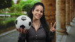 © Krakenimages.com - Woman holding soccer ball and showing peace sign in park with lush greenery and stone columns in background, radiating happiness and enthusiasm outdoors.