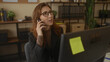 © Krakenimages.com - Woman talking on phone in modern office setting with glasses and computer surrounded by books and files, showcasing a professional business environment indoors.