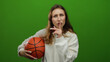© Krakenimages.com - Woman holding basketball making quiet gesture against isolated green background in casual white outfit suggesting calm but focused attitude indoors.