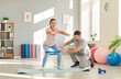 © Studio Romantic - Positive woman in a rehabilitation fitness session with personal trainer in gym. Focused on squats exercise with a band, the male coach guides her workout.