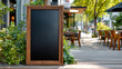 © Anoo - Black notice board in a rustic wooden frame stands prominently outside a restaurant, ready for menu updates, with lush plants and outdoor seating enhancing the scene