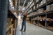 © danr13 - Worker reaching for boxes on high shelves in a large warehouse aisle filled with inventory.