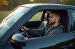 © Pete - Young man with beard drives black car on rural road. He wears grey hoodie, looks forward with calm expression. Outdoors, natural light, autumn foliage in background. Transportation scene.