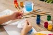 © Andrey_Maksimov - Women's hands wipe the brush with a paper towel to remove excess water after painting. There are different colors of gouache and brushes on the table. Lifestyle. Close-up. Side view.