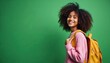 © Viktor - Young african girl with curly hair wears pink sweater and yellow backpack. She smiles looking at camera against green background. Ready for school or travel.