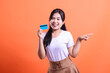 © Ajab - A smiling young woman is holding a credit card, showing her enthusiasm about shopping online. she pointing to the side, isolated on orange background