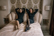 © svetograph - Twins Dresses Bedroom: Identical women pose on bed, indoors, fashion photoshoot.