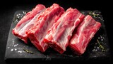 Chunks of raw meat ribs sit on a black stone board. The background is dark. Some salt and herbs surround the meat. The setting is suitable for meal preparation.