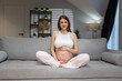 © GRAFStock - Young expectant mother sits cross-legged on a comfortable sofa, cradling her pregnant belly with a gentle smile