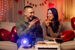 © Home-stock - Romantic evening. Happy European couple eating pizza and watching film on projector, sitting on sofa in room decorated with balloons and heart decorations