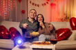© Home-stock - Loving couple having movie night at home, during romantic date, . eating popcorn and pizza while watching film on projector, room filled with red heart balloons for Valentine's Day