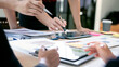 © NAMPIX - Close-up of business people hands pointing at data charts and using a tablet during a strategy meeting in a bright, modern office space.