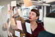 © TrueFrame Collective - Caucasian young adult woman brainstorming ideas with colleagues, writing on transparent board covered with sticky notes, collaborating in modern office environment during meeting