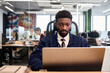 © TrueFrame Collective - Young adult Black man sitting at desk working on laptop in modern open plan office, focused expression, other people collaborating in background, glass partition visible