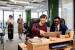 © TrueFrame Collective - Caucasian young adult man and Black young adult man sitting at desk collaborating on laptop in modern office while two young adult women standing in background