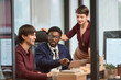 © TrueFrame Collective - Young adult Caucasian man, young adult Black man, young adult Caucasian woman collaborating in modern office, discussing project at desk with laptop and documents, working together
