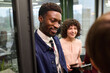 © TrueFrame Collective - Young Black man smiling while standing near glass door, accompanied by young Caucasian woman holding digital tablet, both engaging in conversation with unseen colleague in office setting