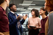 © TrueFrame Collective - Group of young adult multiethnic colleagues standing together in modern office, Black man gesturing while talking, Caucasian woman holding digital tablet, others listening and smiling
